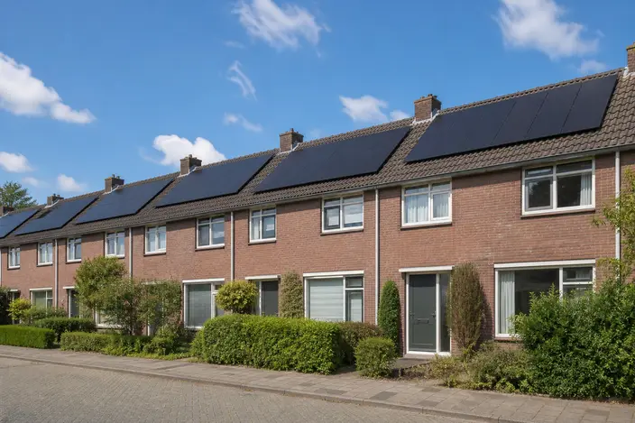 AI-beeld: een rij Nederlandse rijtjeshuizen van rode baksteen met donkere zonnepanelen op de schuine daken, onder een blauwe lucht
